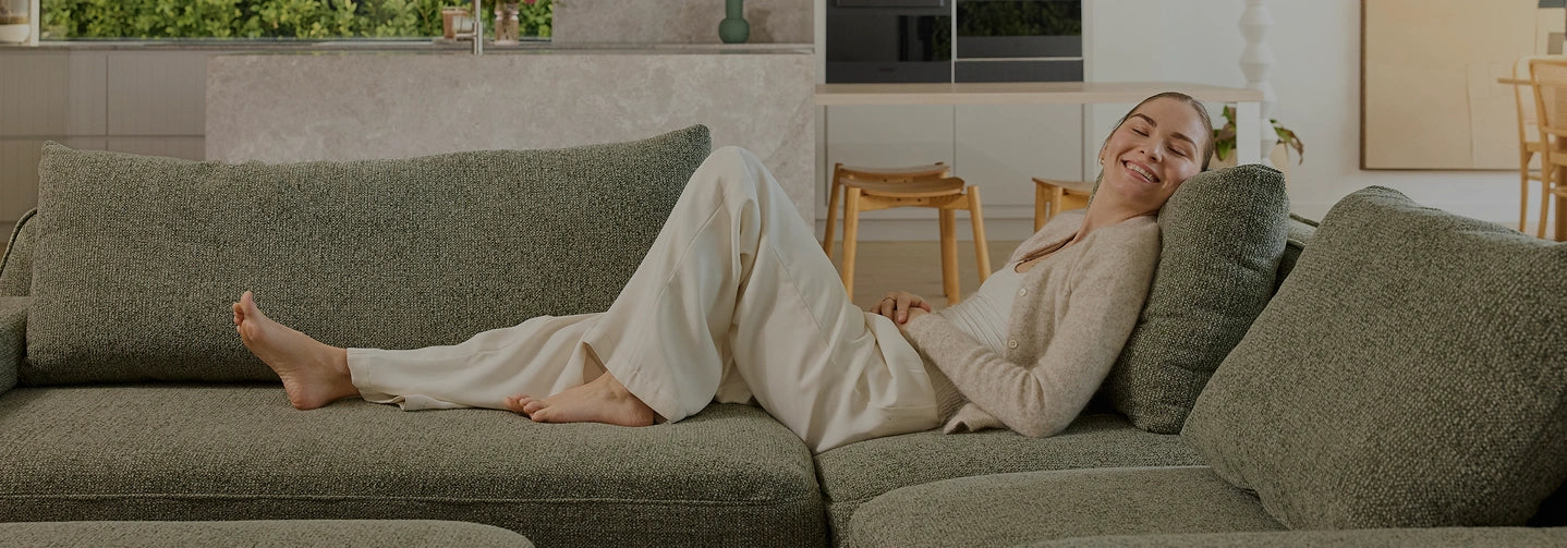 A woman in a beige lounge set reclines comfortably on a textured gray-green sectional sofa in a modern living room setting.