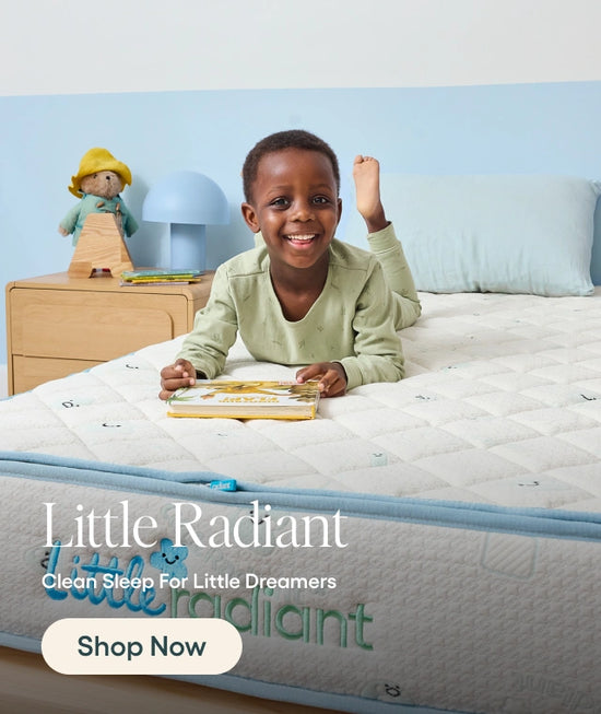 A smiling young child lies on a quilted mattress in a bright bedroom setting, with a light blue wall, bedside table, lamp, and doll in the background. Large text at the bottom reads 'Little Radiant' and 'Clean Sleep For Little Dreamers' with a 'Shop Now' button.