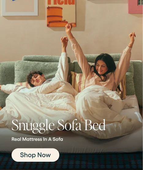 A lifestyle advertisement showing a man and a woman waking up together on a green upholstered sofa bed, covered in white bedding. The text overlay reads 'Snuggle Sofa Bed', 'Real Mattress In A Sofa', and 'Shop Now'.