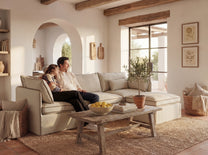 Living room with a couple sitting on a beige sofa, a wooden coffee table with fruit bowl and books, and decorative elements.