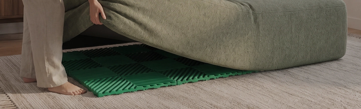 A person in light beige pants is lifting a sage green upholstered ottoman or bench to reveal a bright green textured floor mat underneath, set on a light wood floor.
