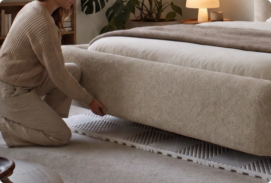 A woman kneels beside a bed in a softly lit bedroom, adjusting a textured beige upholstered bed frame or sofa base placed over white slatted floor mats. The room has neutral tones, plants, and a lamp in the background.