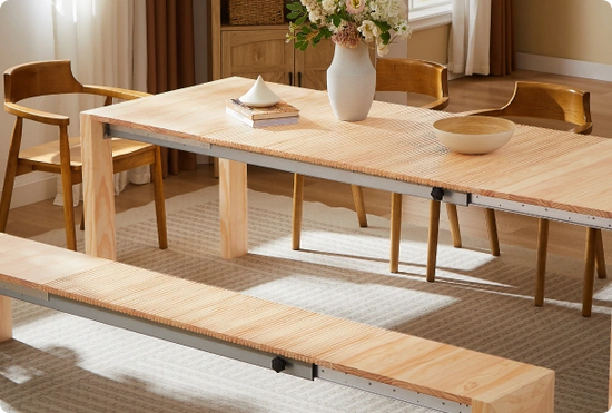 A light natural wood dining table set in a bright room, with matching wooden chairs and a bench, styled with a white vase of flowers and a bowl.