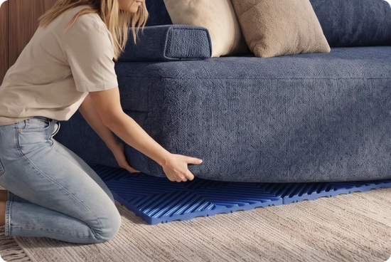 A woman kneels beside a blue upholstered sofa on a textured beige rug, placing a blue slatted floor mat or pad underneath the sofa.