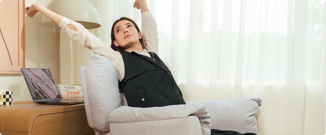 Woman sitting on a chair with arms raised, wearing a black jacket and beige sweater, in a room with a desk and laptop.