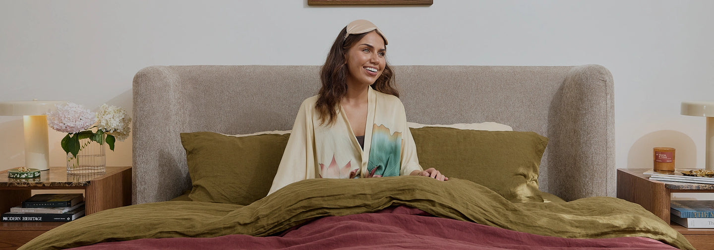 A woman sits in bed wearing a cream robe and a matching headband, surrounded by olive green pillows and a burgundy blanket in a softly lit bedroom.