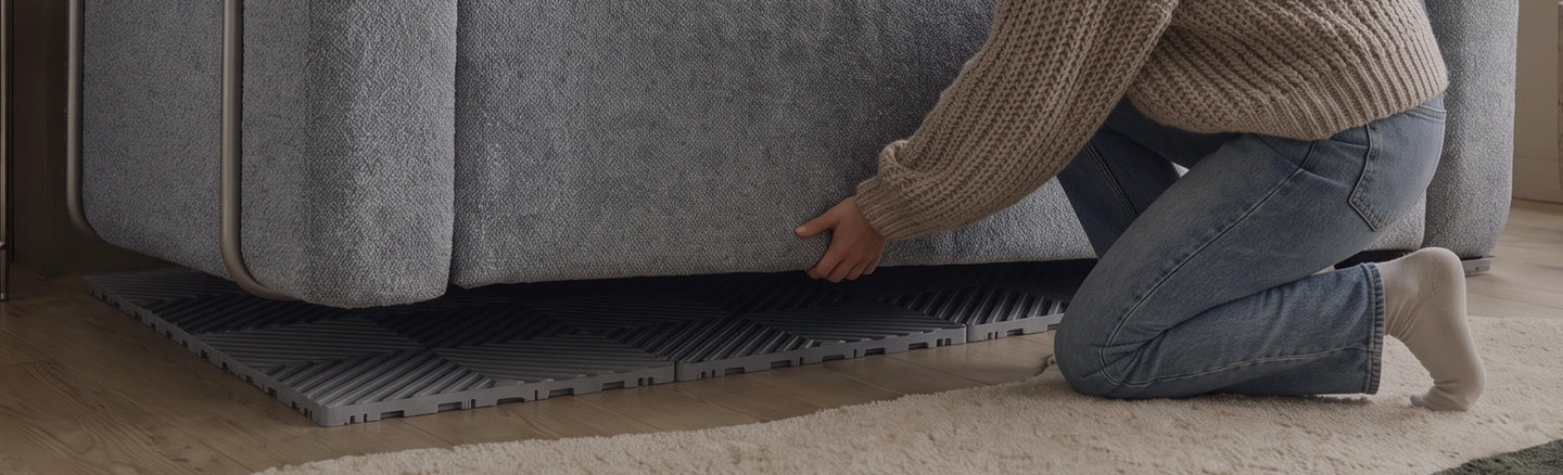 A person kneels under a gray upholstered sofa to place or adjust a dark gray textured floor mat or rug on the floor.