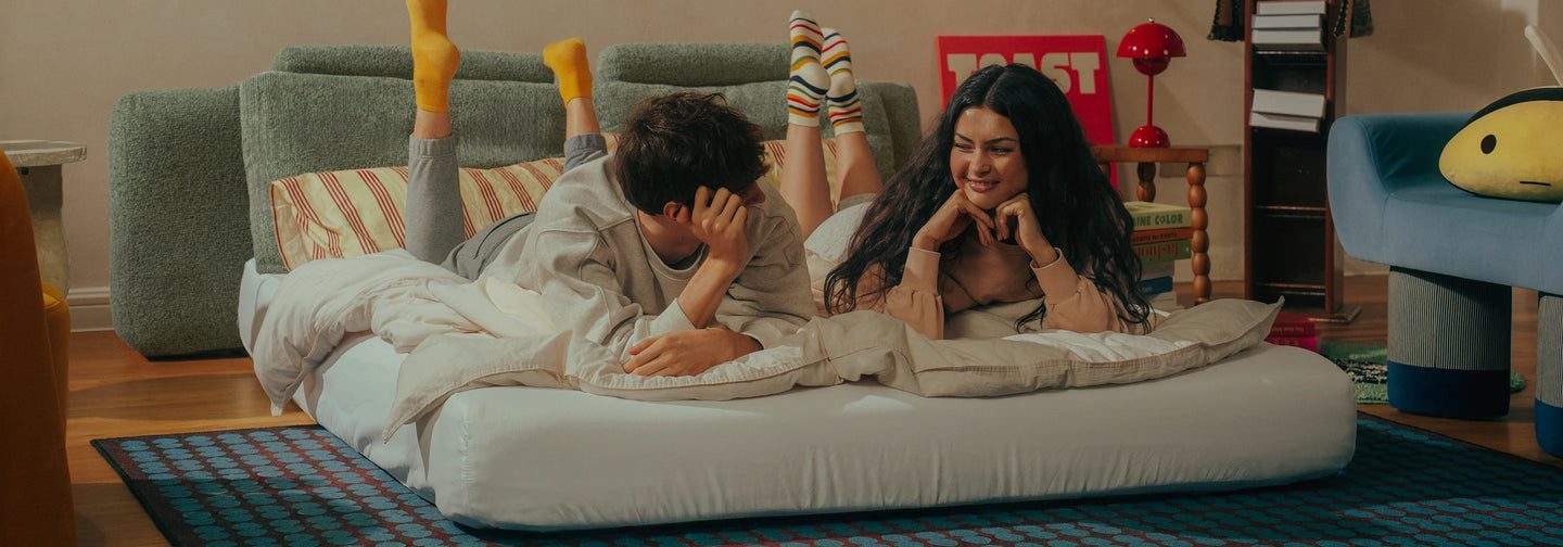 A man and a woman lying on a white mattress on the floor of a living room, with their legs raised in the air. The man wears a light gray sweatshirt and bright yellow socks, while the woman wears a white top and striped socks.