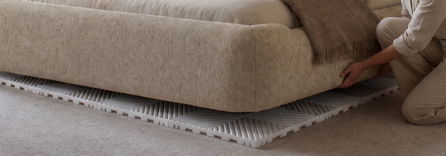 A person kneels beside a beige upholstered sofa to place or adjust a white textured floor mat or rug underneath the sofa in a living room setting.