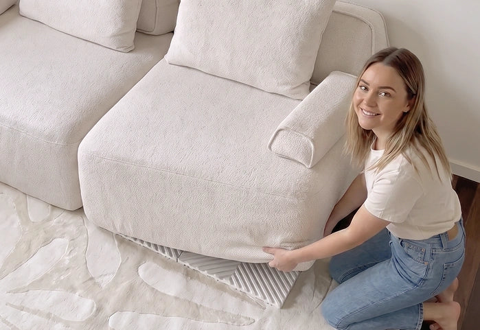 A woman kneels beside a chunky, off-white bouclé sofa, adjusting or showing the underside/base of the seat cushion. The scene is a bright, modern living room with a matching sofa, white rug, and light wall.