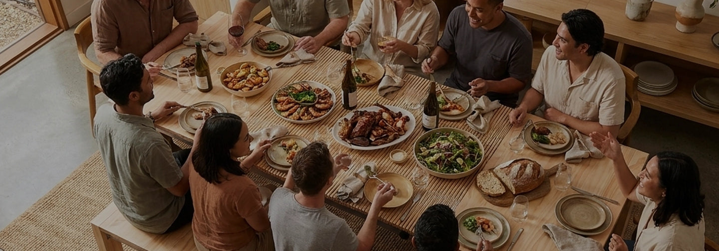 A group of people gathered around a wooden dining table, sharing a large feast with plates of food, bottles, and glasses in a warm, rustic indoor setting.